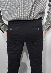 Pantaloni neri con una texture liscia, dotati di due tasche posteriori e dettagli con bottoni. Abbinati a una camicia a maniche lunghe fantasia.