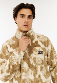 Fleecepullover i beige og krem med et mønstret tekstur. Har en høj krave, halv lynlås og en brystlomme med logo.