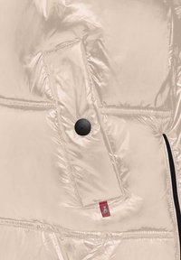 Zbliżenie na beżową pikowaną kieszeń kurtki z czarnym zatrzaskiem i małą czerwoną metką w pobliżu szwu zamka.