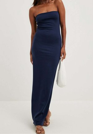 Femme portant une robe maxi bleu marine sans bretelles, tenant un sac à main blanc et chaussée de sandales couleur beige sur un fond clair uni.