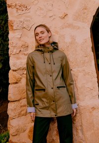 Donna con impermeabile verde oliva con tasche con cerniera nera, in piedi rilassata contro un muro di pietra testurizzato con un'apertura ad arco.