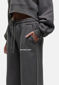 Grauer, verkürzter Sweatshirt und passende Jogginghose mit elastischem Bund. Die Hose hat Seitentaschen und weißen Logo-Text auf dem Oberschenkel.