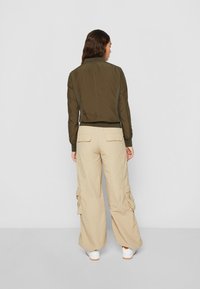 Oliven grønn bomberjakke med ribbet krage og mansjetter, sammen med beige cargo bukser med sidelommer og en løs passform. Hvite sneakers er synlige.