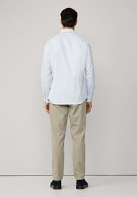 Muškarac stoji okrenut leđima u svijetloplavoj kariranoj košulji dugih rukava, bež hlačama i tamnim cipelama, nasuprot jednostavnoj pozadini.