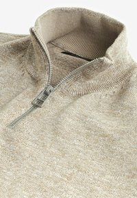 Beżowy sweter z dzianiny z wysokim kołnierzem i zamkiem na pół. Posiada fakturowany wzór i subtelne logo wzdłuż brzegu.