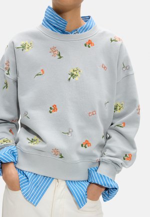 Osoba ubrana w jasnoszarą bluzę z małymi, kolorowymi, haftowanymi kwiatami, pod którą ma niebiesko-białą koszulę w paski, z rękami w kieszeniach.