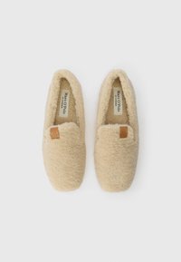 Par bežastih krznenih papučica s kvadratnim prstima i malim smešnim kožnim oznakama na vrhu, označenim imenom Marc O'Polo unutra.