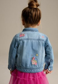 Hellblaues Denim-Jackett mit gestickten Tier-Patches und floralem Muster auf dem Rücken. Die Ärmel sind mit Knöpfen versehen. Eine rosa Tüllröcke ist sichtbar.