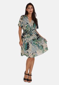 Bloemrijke wikkeljurk in lichtbeige met groene en blauwe patronen. Korte pofmouwen en een elastische taille. Gecombineerd met zwarte haksandalen.