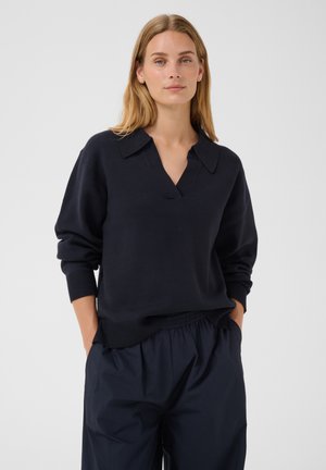 Femme aux cheveux blonds portant un pull à manches longues bleu marine foncé avec un col et un pantalon ample bleu marine foncé, mains dans les poches, fond neutre.