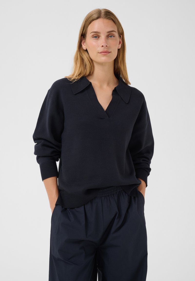 Femme aux cheveux blonds portant un pull à manches longues bleu marine foncé avec un col et un pantalon ample bleu marine foncé, mains dans les poches, fond neutre.