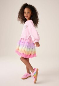 Ragazza con capelli ricci che indossa un maglione rosa con una toppa a forma di cuore, gonna a strati color arcobaleno e scarpe da ginnastica rosa con strisce arcobaleno, in piedi di lato.