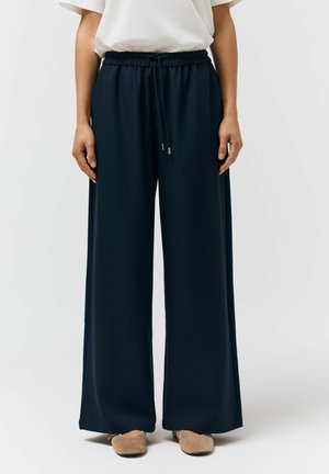 Osoba nosząca luźne granatowe szerokie spodnie z elastycznym pasem i sznurkiem, w połączeniu z beżowymi płaskimi butami i białą bluzką.