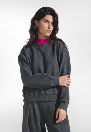 Kvinde med mørkt hår iført en grå fiskebensmønstret sweatshirt og bukser med en lyserød rullekrave, der kigger let til siden.