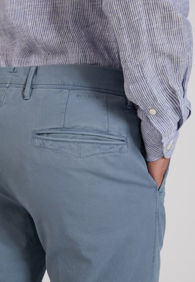 Man wearing blue trousers with back pocket and striped long-sleeve shirt, hand resting in side pocket.