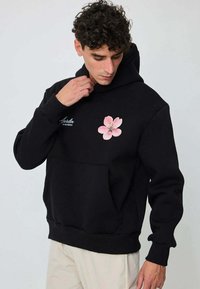 Homme portant un sweat à capuche noir avec un motif de fleur rose et le texte « Jardin de Provence », ajustant le col, debout devant un fond uni.