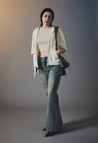 En modell bærer en cropped hvit hettegenser, beige topp og stripete blå utsvingte bukser, med en grønn veske og en protesebelagt venstre arm. Sorte sko fullfører antrekket.
