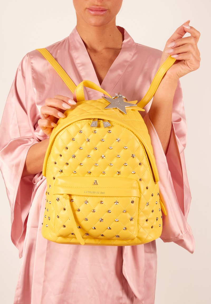 Geel gewatteerde rugzak met zilveren steraccenten en hardware. Beschikt over een voorkantvak en verstelbare schouderbanden. Glad, glanzend oppervlak.