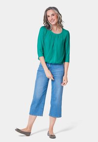 Groene blouse met korte mouwen en een geplooide voorkant, gecombineerd met lichtblauwe cropped jeans en grijze ballerina's, tegen een witte achtergrond.