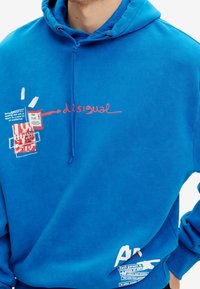 Modrá mikina s kapucí s předním grafickým motivem obsahujícím text a abstraktní tvary. Materiál je měkký, s volným střihem a kapucí na stahovací šňůrku.