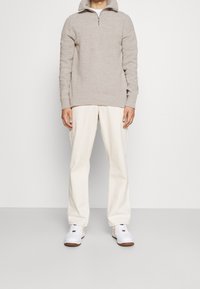 Pull en tricot beige avec un col zippé à moitié, associé à un pantalon large couleur crème et des sneakers blanches avec des accents marron.