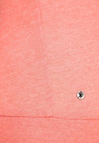 Korálově růžová mikina s jemnou texturou, s malým černým logem na boku a žebrovaným lemem.