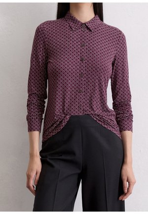 Femme portant une chemise bordeaux ajustée à manches longues avec un motif géométrique blanc et un pantalon noir taille haute.