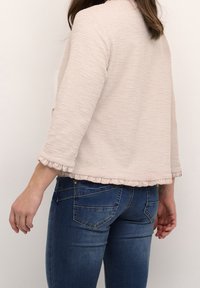Woman wearing light pink textured three-quarter sleeve top with ruffle edge and blue denim jeans, standing against white background.