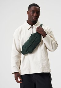 Dunkelgrüne, stoffliche Umhängetasche, die über die Brust getragen wird, mit zwei farbigen, runden Akzenten und einem Reißverschluss. Mann in neutralem Gesichtsausdruck.