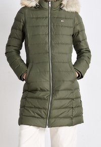 Olivgrüner Puffer-Mantel mit Kapuze aus Kunstpelz, gestepptem Design, Reißverschluss und seitlichen Taschen. Mit einem Logopatch auf der oberen Brust.