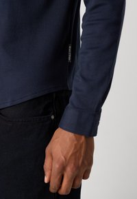 Marineblå langt skjorte laget av et mykt stoff, med rundet kant og en merkelapp på siden. Bært med mørk denimbukser.