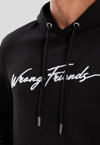 Sudadera negra con cordones, que presenta "Wrong Friends" bordado en blanco. Tejido suave, diseño casual y detalles metálicos en los extremos de los cordones.