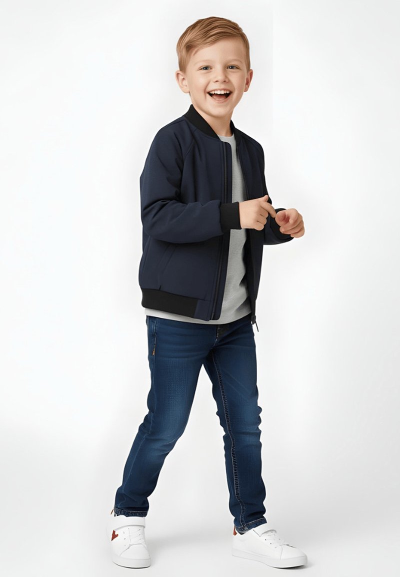 Granatowa kurtka bomberka ze ściągaczami na mankietach i u dołu, szara koszulka, ciemnoniebieskie jeansy oraz białe sneakersy. Dziecko uśmiechnięte, stojące.
