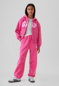 Mladi oseba stoji z rokami v žepih, oblečena v svetlo roza flis s kapuco in hlačami, bele superge in belo majico.