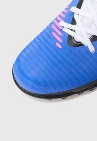 Μπλε ποδοσφαιρικό παπούτσι με υφασμένο συνθετικό επάνω μέρος, μαύρη σόλα, ροζ λεπτομέρειες και λευκά κορδόνια. Διαθέτει ραβδώσεις για καλύτερη πρόσφυση και έλεγχο.