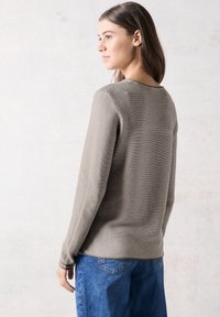 Maglione a maniche lunghe color taupe in costina con scollatura rotonda, abbinato a jeans in denim blu a gamba larga. La texture è morbida; è visibile una leggera elasticità.
