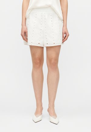 Mujer vistiendo shorts blancos de talle alto con patrones verticales de ojales y tacones puntiagudos blancos con detalles de encaje, posando frente a un fondo liso.