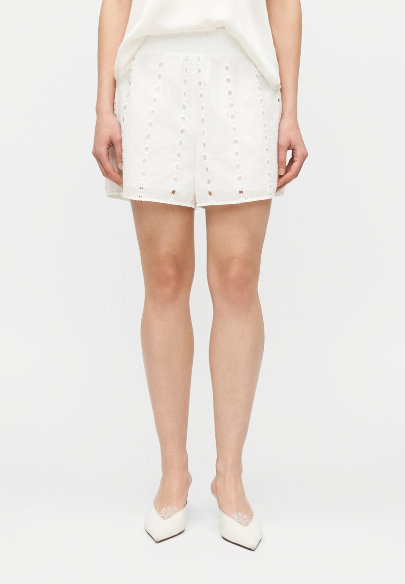 Γυναίκα που φοράει λευκά σορτς ψηλού καβάλου με κατακόρυφα σχέδια από κεντήματα και λευκές μυτερές γόβες με λεπτομέρειες δαντέλας, όρθια μπροστά σε απλό φόντο.