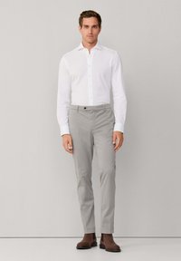 Mężczyzna stojący w białej koszuli zapinanej na guziki, jasnoszarych spodniach i brązowych botkach typu Chelsea na jednolitym jasnoszarym tle.