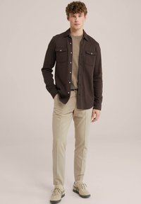 Brown button-up shirt with two chest pockets, layered over a beige crew-neck sweater; paired with light beige trousers and beige sneakers.