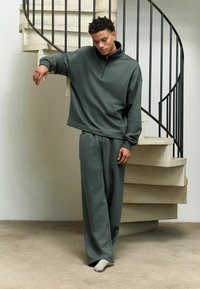 Sudadera de color verde oscuro con cuello alto y cremallera de un cuarto, combinada con pantalones de chándal holgados a juego. Tejido suave y corte relajado.