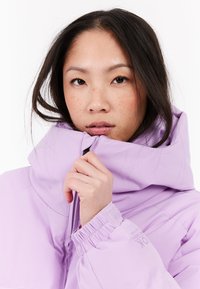 Mladá žena s dlhými tmavými vlasmi, oblečená v svetlofialovej pérovke, drží zips blízko brady na bielom pozadí.