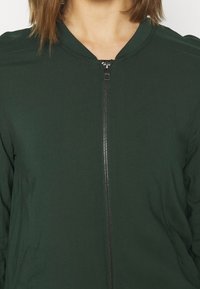 Veste bomber vert foncé en tissu léger, dotée d'une fermeture éclair et d'un col arrondi. Comprend des poches latérales et une texture lisse.