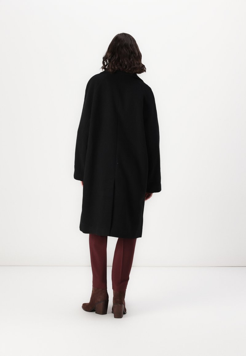 Schwarze Jacke mit lässigem Schnitt, ausgestattet mit langen Ärmeln und einer Naht auf der Rückseite, kombiniert mit bordeauxfarbenen Hosen und braunen Absatzstiefeln.