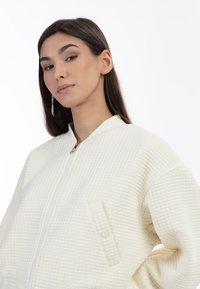 Witte gewatteerde ritsjack met een hoge kraag en een drukknopsluiting op de patch pocket aan de voorkant. Gladde textuur en subtiele patroon details.