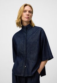 Camicia a maniche corte in denim scuro con colletto rialzato, chiusura con bottoni frontale e taschino sulla parte sinistra. Tessuto liscio e vestibilità comoda.