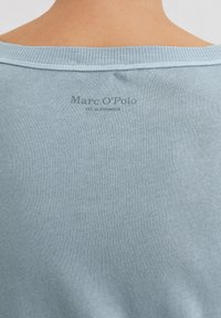 Hellblauer Pullover mit geripptem Ausschnitt, weichem Material und bedrucktem Logo "Marc O'Polo" auf der Rückseite. Einfaches, lässiges Design.