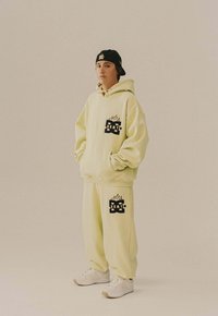 Joven vestido con un hoodie de gran tamaño de color amarillo claro y pantalones de chándal con logotipos gráficos en negro, zapatillas blancas y una gorra negra puesta al revés.