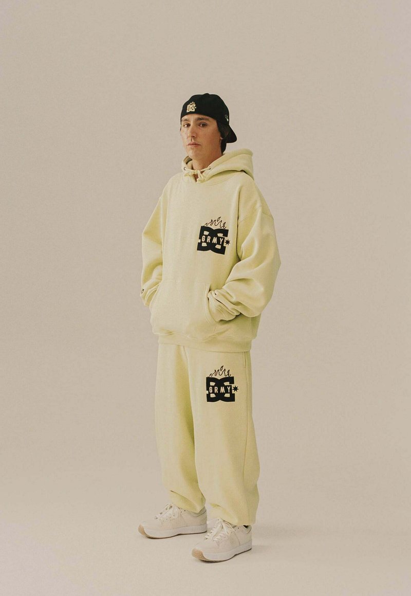 Joven vestido con un hoodie de gran tamaño de color amarillo claro y pantalones de chándal con logotipos gráficos en negro, zapatillas blancas y una gorra negra puesta al revés.