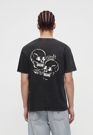 Person with braided hair wearing a black t-shirt with white skulls and text "Replay Since 1981" on the back, paired with light blue jeans.